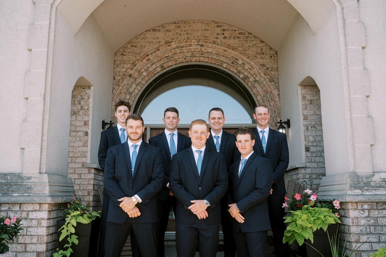 Seven men in dark suits and blue ties posing in front of a brick arched house entrance with potted flowers.