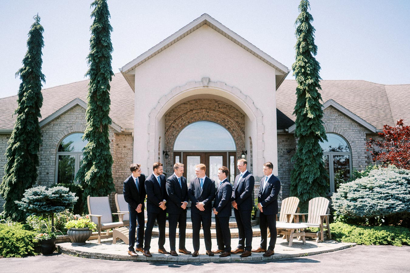 Groom and groomsmen in navy suits standing outside a large family home with arched entrance, evergreen trees, garden shrubs, and patio chairs.
