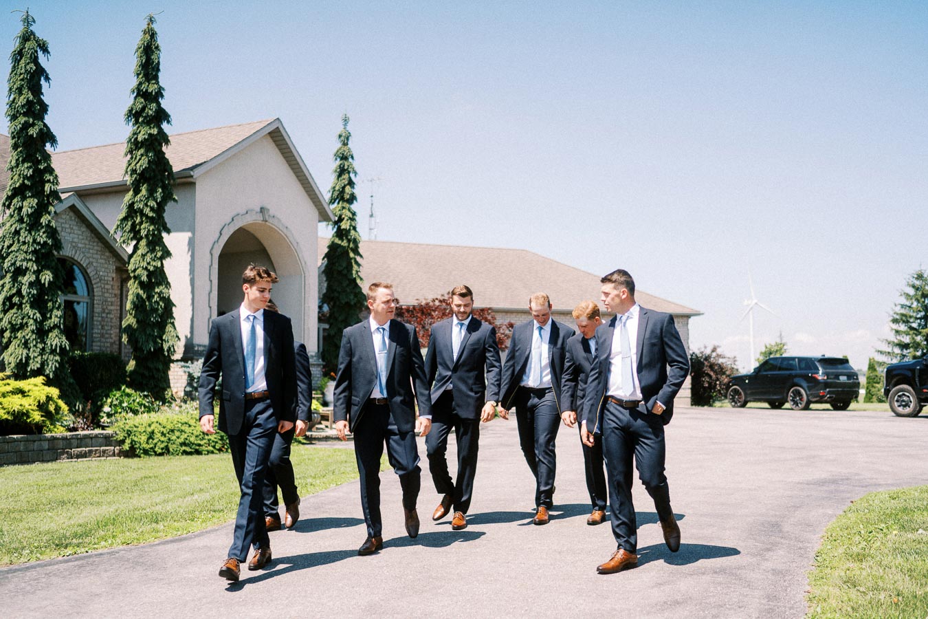 Groom and groomsmen in navy suits walking on a driveway outside a rural family home near Chatham-Kent, Ontario, Canada.