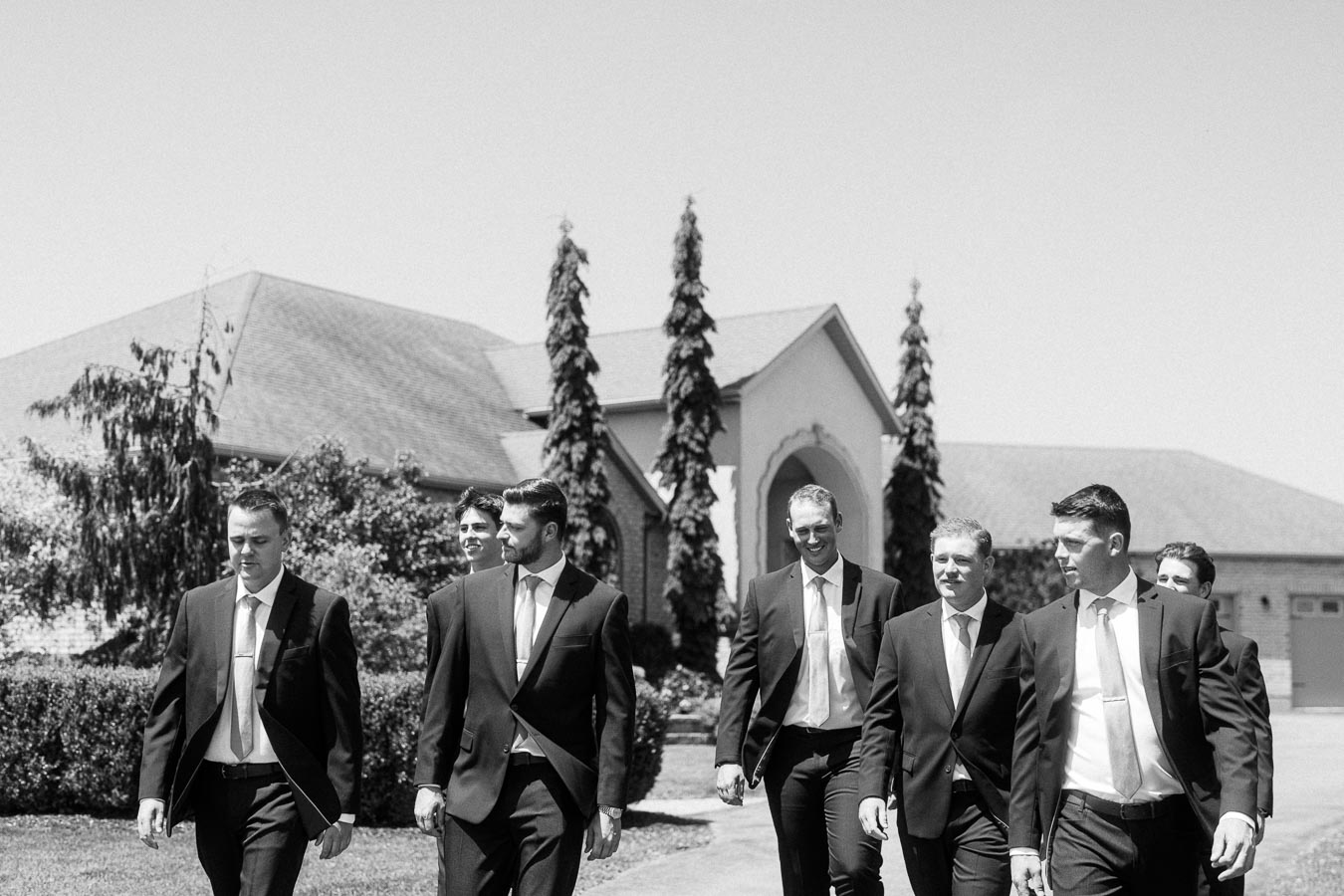 Black and white group portrait of groom and groomsmen in suits walking outside a family home in Chatham-Kent, Ontario, Canada.
