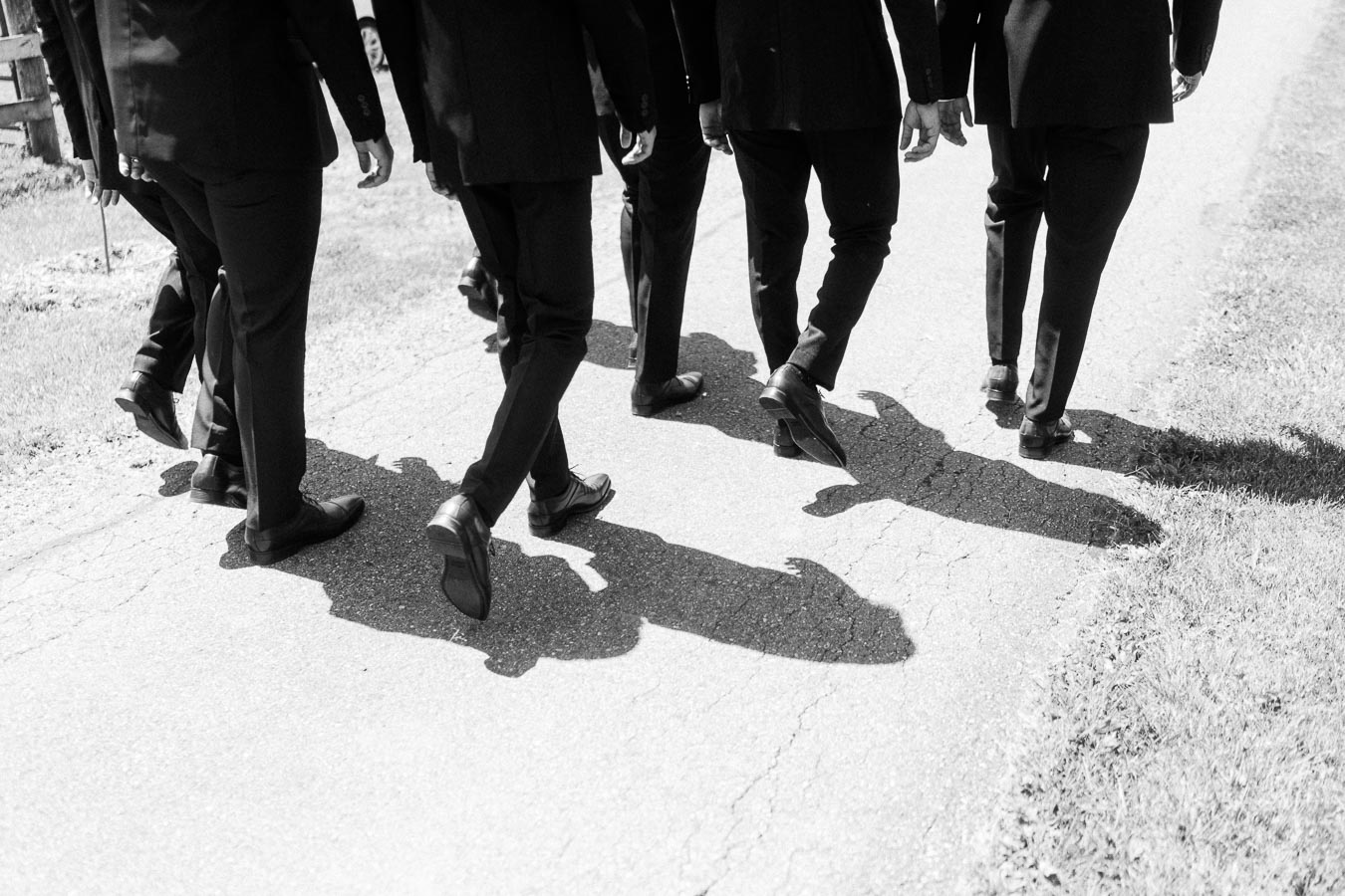 Black and white photo of groom and groomsmen in suits walking on a rural driveway with long shadows on a sunny day.
