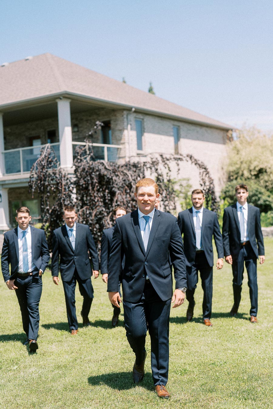 Groom and groomsmen in navy suits walking across a lawn in front of a family farmhouse near Chatham-Kent, Ontario.
