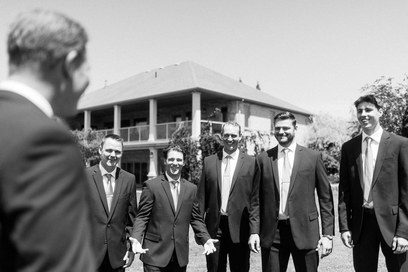Black and white outdoor group portrait of groom and groomsmen in suits smiling in front of a rural family house.