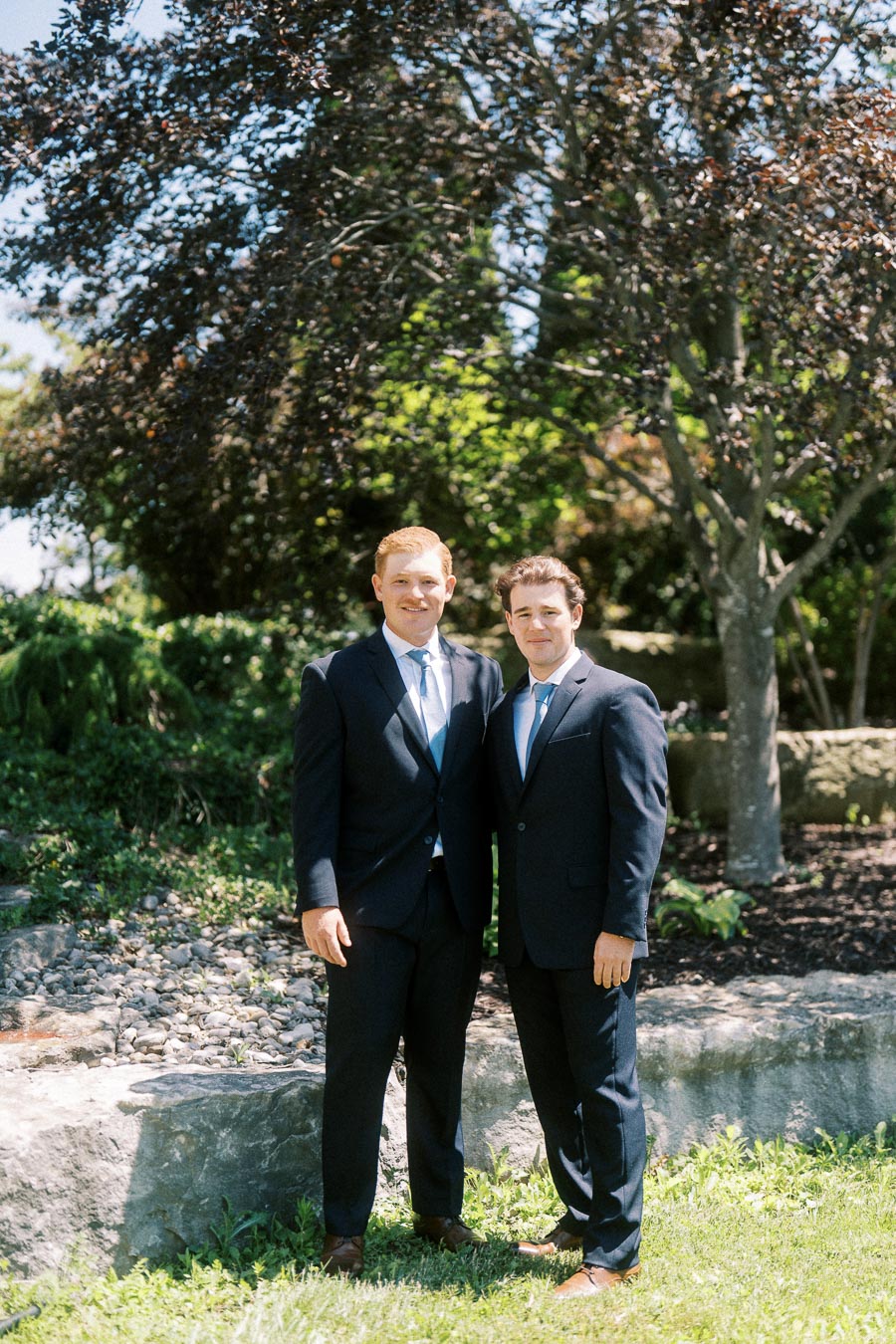 Two men in dark suits pose together outdoors in a sunny garden with trees, grass, stones, and a low rock wall.