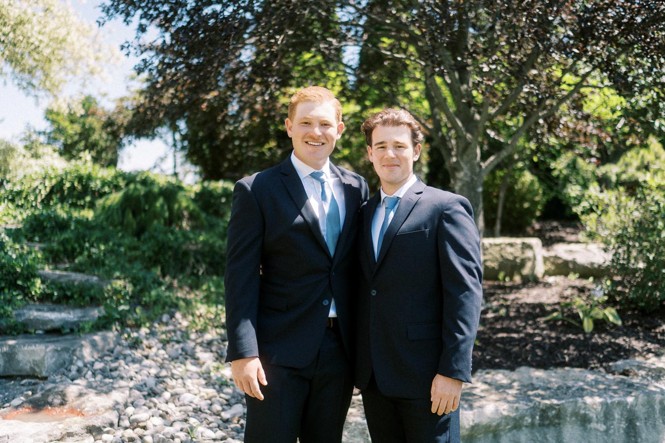 Two men in dark suits and blue ties posing together in a sunny garden with trees, rocks, and greenery.