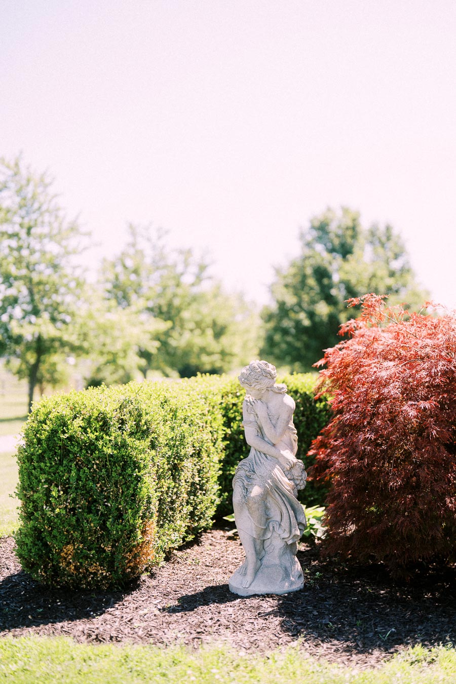 Garden statue of a woman among manicured hedges and red foliage in bright sunlight.