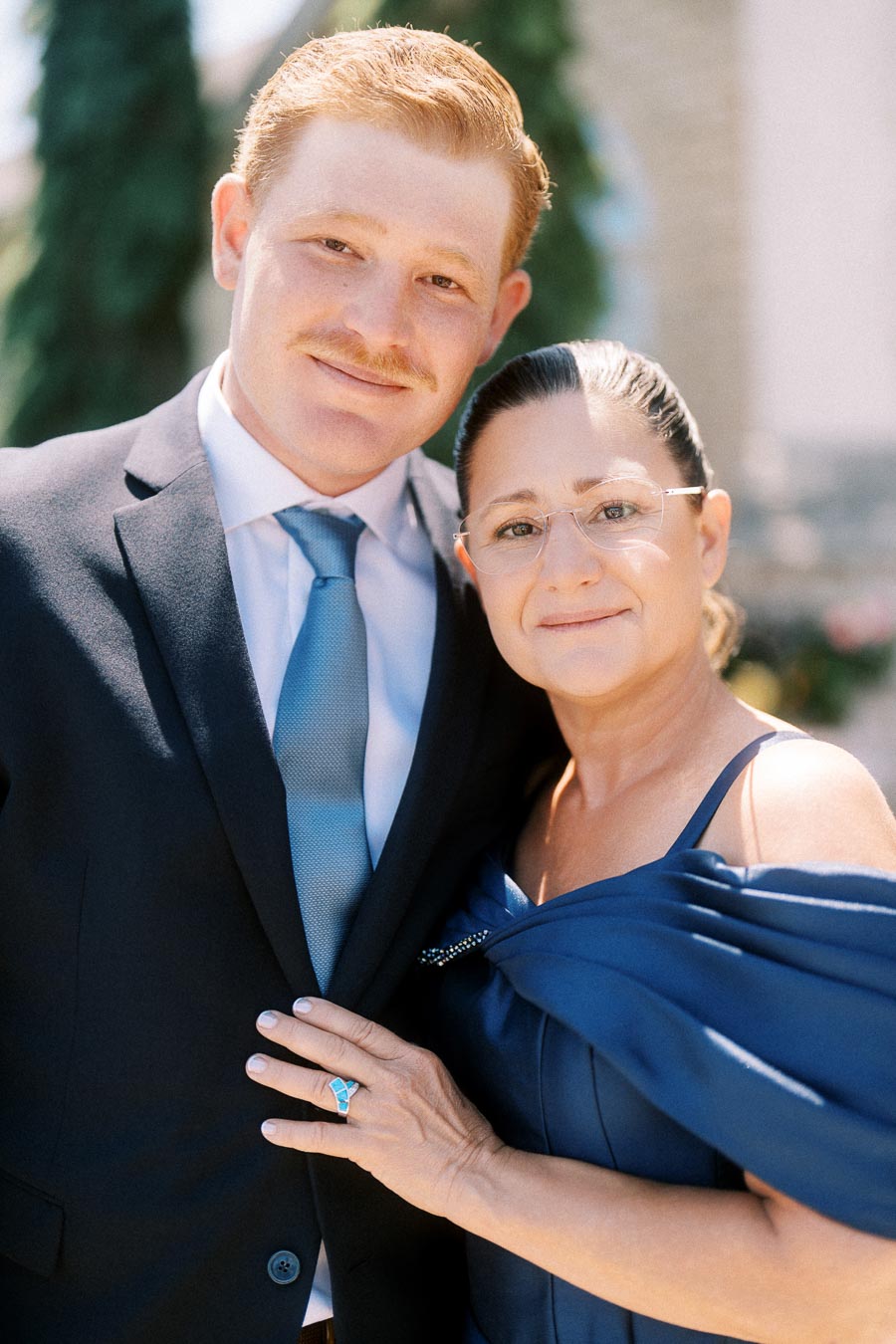 Groom in a black suit and blue tie posing with his mother in a navy dress outdoors near Chatham-Kent, Ontario before a wedding.
