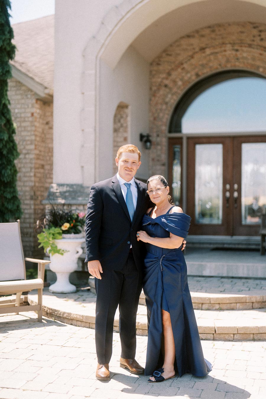 Groom in navy suit posing with his mother in a blue dress outside a brick family home near Chatham-Kent, Ontario, Canada before a wedding.