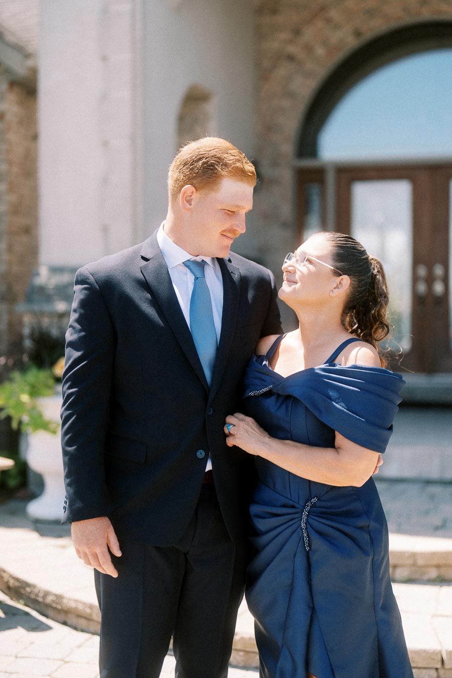 Groom in a navy suit posing with his mother in a blue dress outside a brick home near Chatham-Kent, Ontario.