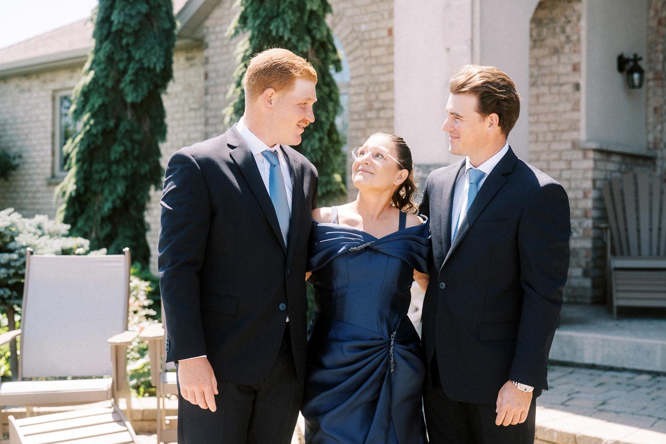 Two groomsmen in navy suits stand with a smiling woman in a navy dress outside a brick family home in Chatham-Kent, Ontario.