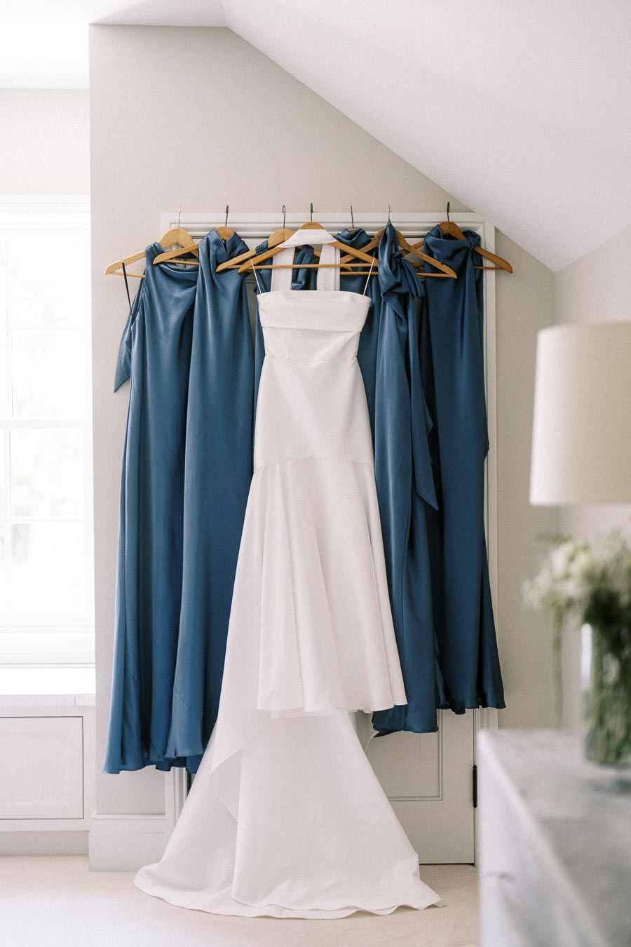 White strapless wedding dress hanging with blue bridesmaid dresses in a bright room before an Ontario farm wedding.