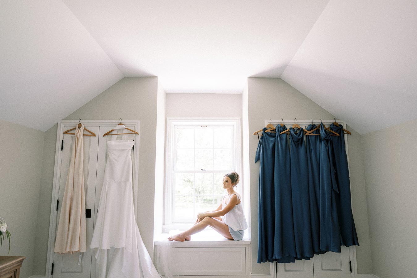 Bride sitting on a bright window seat between hanging wedding dress and blue bridesmaid dresses during morning preparations in Chatham-Kent, Ontario.