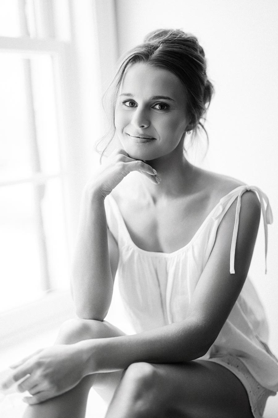 Black and white portrait of a smiling bride sitting by a bright window during wedding morning preparations in Chatham-Kent, Ontario.