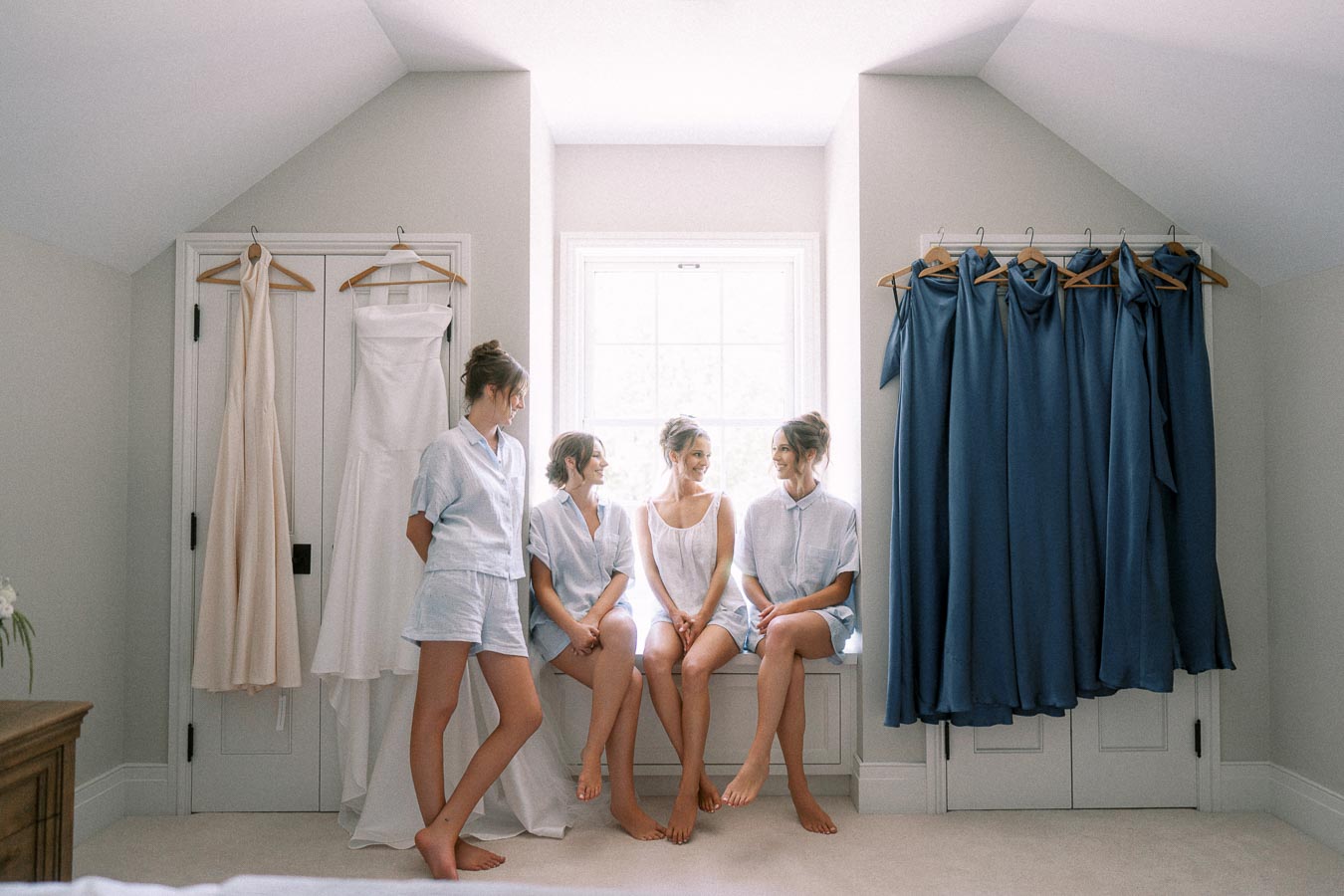 Bride and bridesmaids in light blue pajamas sit by a bright window between hanging wedding dress and blue bridesmaid dresses in Ontario farmhouse bedroom.