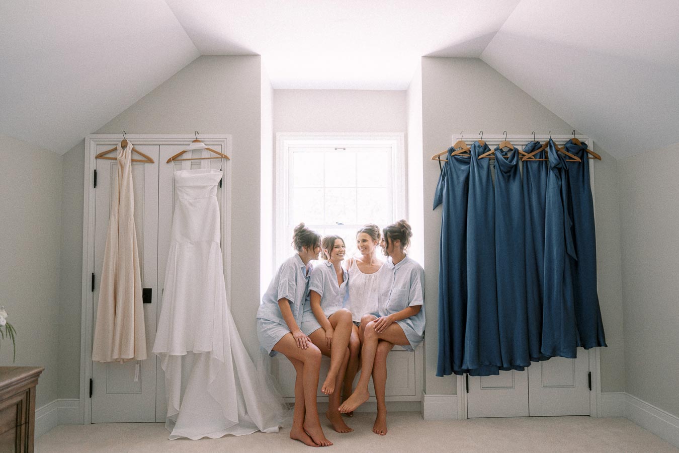 Bride and bridesmaids sit by a bright window with wedding dress, veil, and blue bridesmaid dresses hanging in an Ontario farmhouse room.