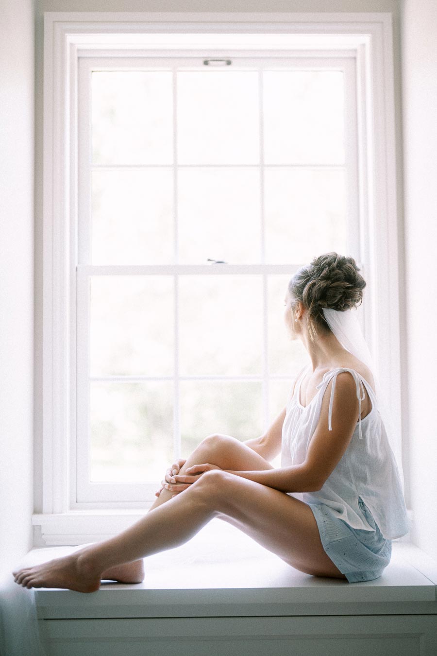Bride sitting on a white window seat in soft morning light before an Ontario farm wedding near Chatham-Kent.