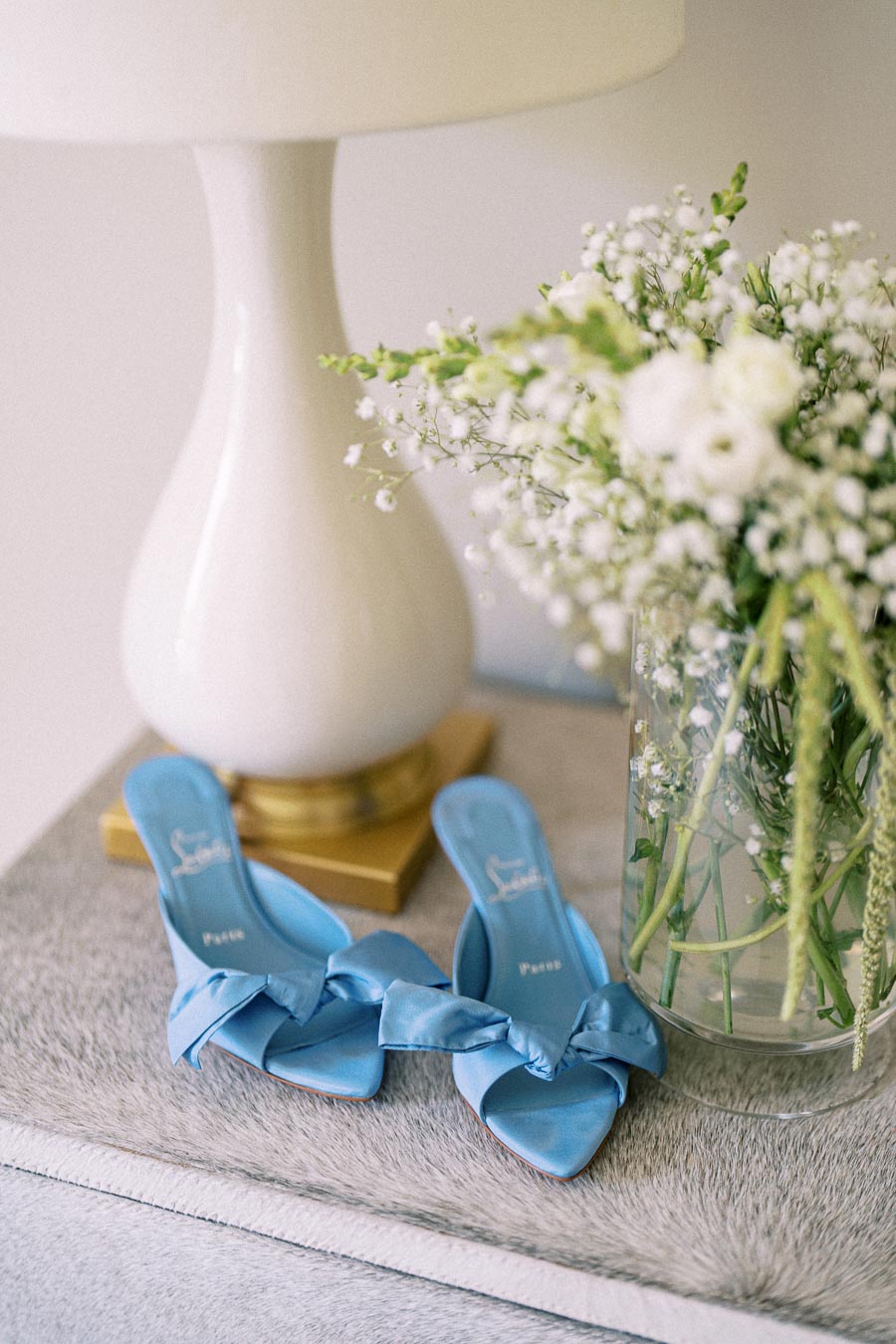 Blue bridal heels with bow details beside a glass vase of white flowers and a white table lamp on a soft gray surface.