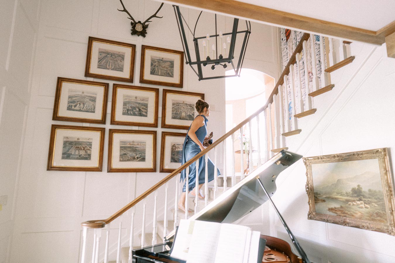 Woman in blue dress walking upstairs in a bright farmhouse interior with framed art, chandelier, staircase, and grand piano.