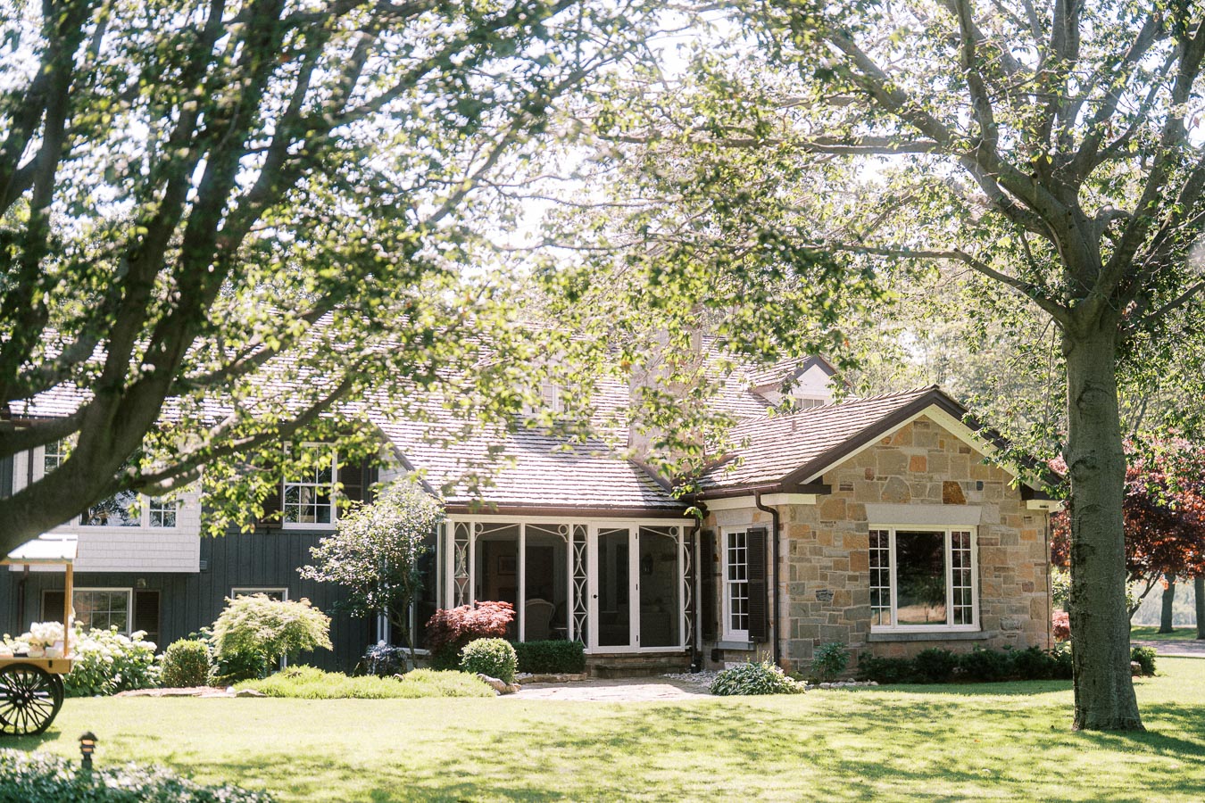 Stone farmhouse on a sunny private farm estate lawn in Chatham-Kent, Ontario, with mature trees, garden shrubs, porch, and rustic wagon.