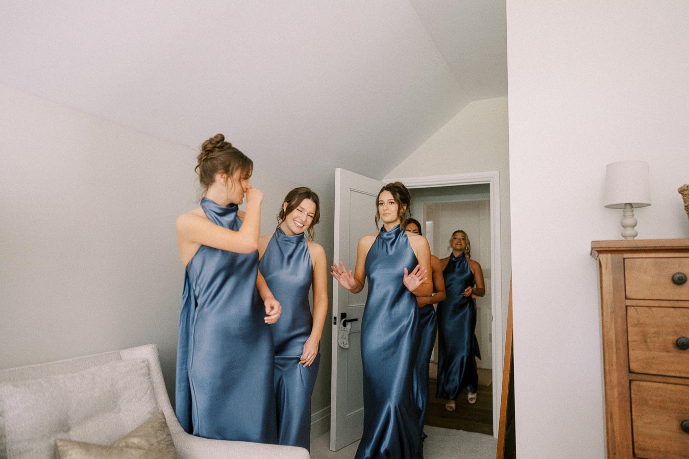 Bridesmaids in blue satin dresses entering a bright farmhouse bedroom during wedding morning preparations near Chatham-Kent, Ontario.