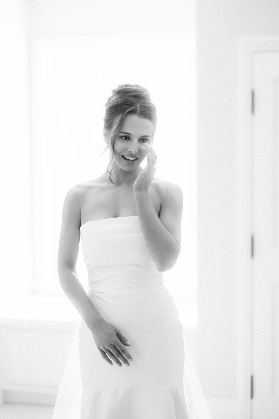 Black and white portrait of a bride in a strapless wedding dress standing indoors by a bright window in Chatham-Kent, Ontario.