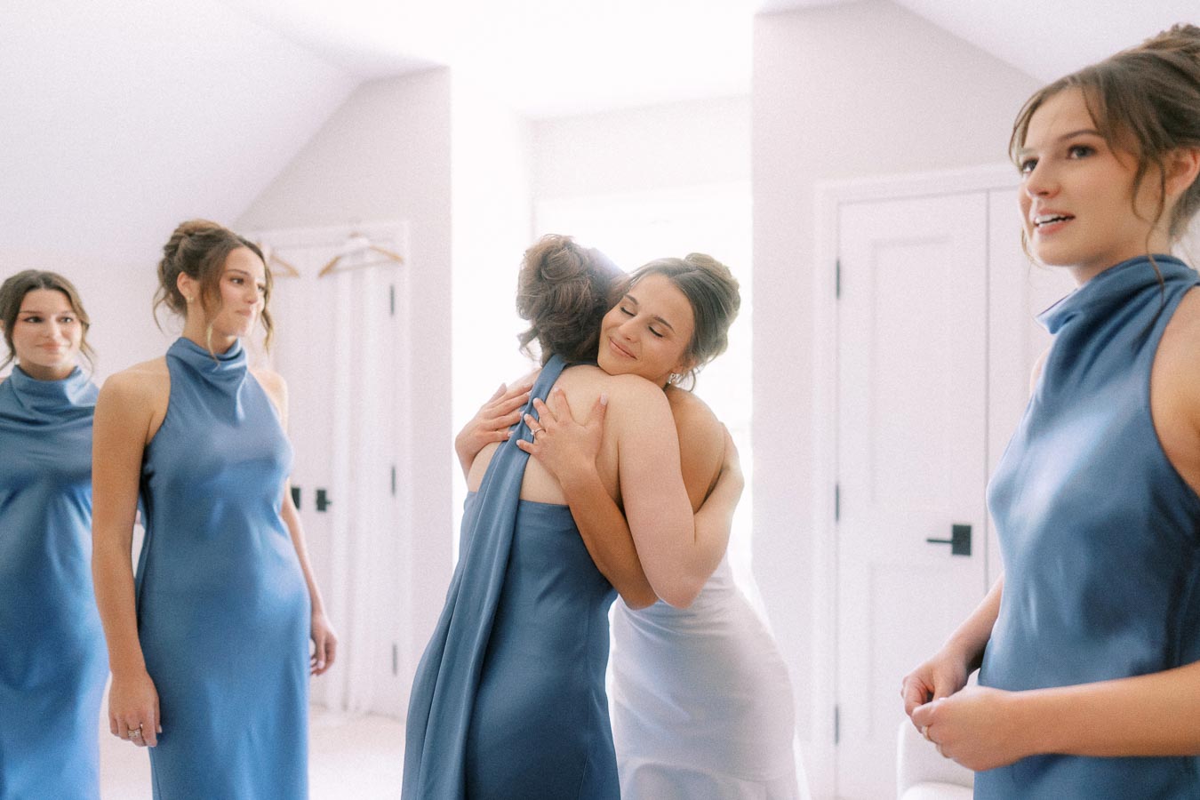 Bride in a white dress hugs a bridesmaid in a blue satin dress during wedding morning preparations in a bright room near Chatham Kent, Ontario.