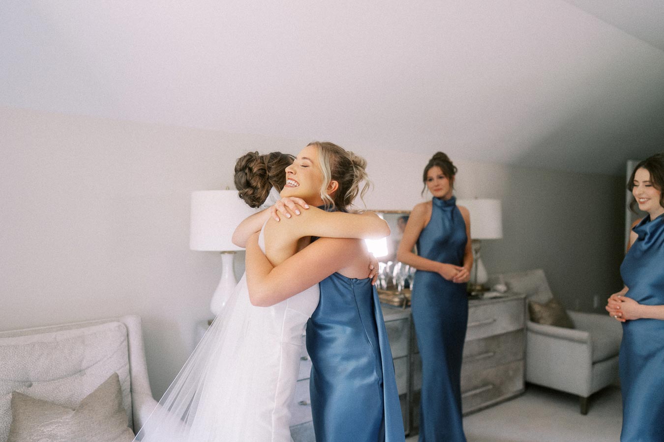 Bride in a white gown and veil hugs a bridesmaid in a blue dress while other bridesmaids watch in a bright room near Chatham-Kent, Ontario.