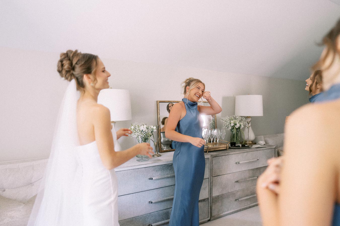 Bride in a white dress and veil laughs with bridesmaids in blue dresses during wedding morning preparations in a bedroom near Chatham-Kent, Ontario.