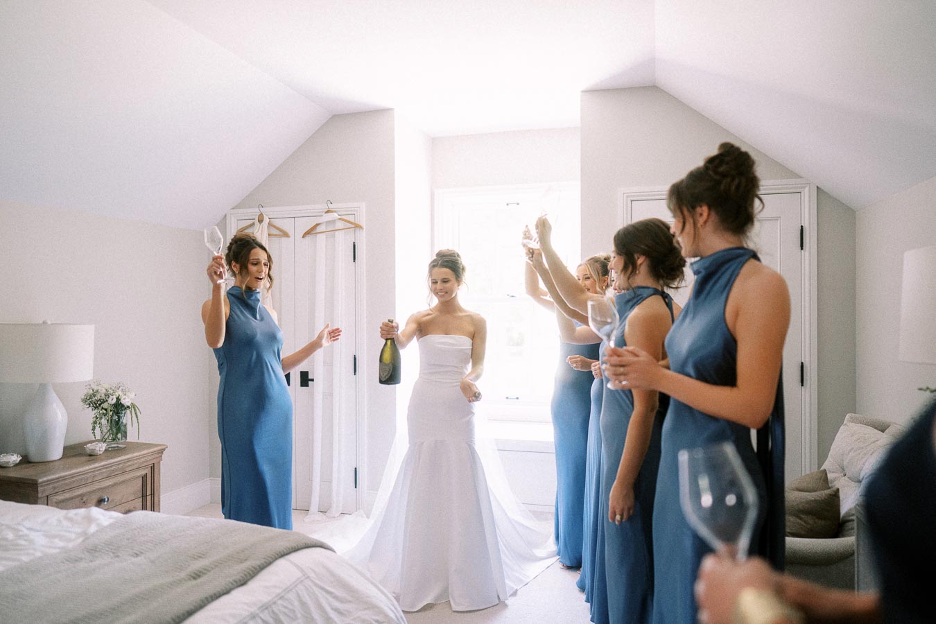 Bride in a white gown celebrates with bridesmaids in blue dresses holding champagne glasses in a bright bedroom near Chatham-Kent, Ontario.