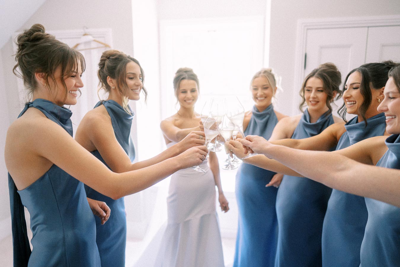 Bride and bridesmaids in blue dresses raising champagne glasses during wedding morning preparations near Chatham-Kent, Ontario.
