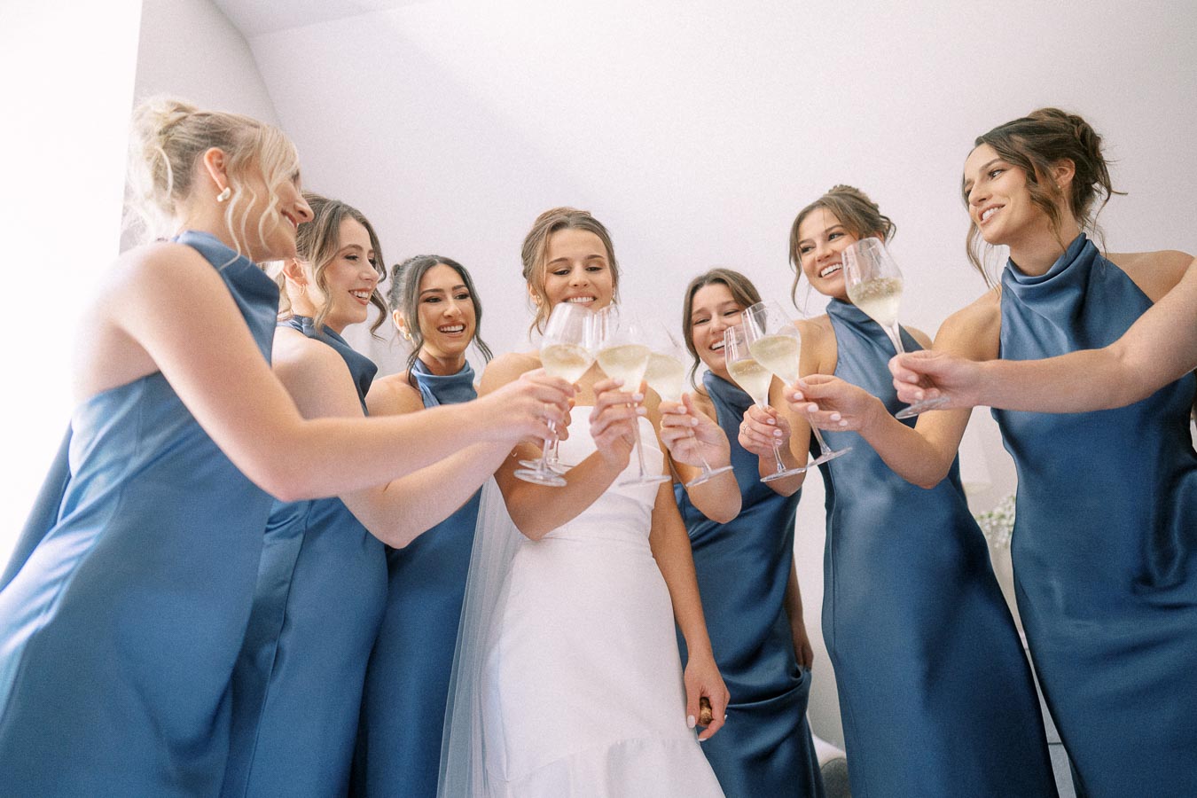 Bride and bridesmaids in blue dresses toast with champagne glasses during wedding morning preparations near Chatham-Kent, Ontario, Canada.