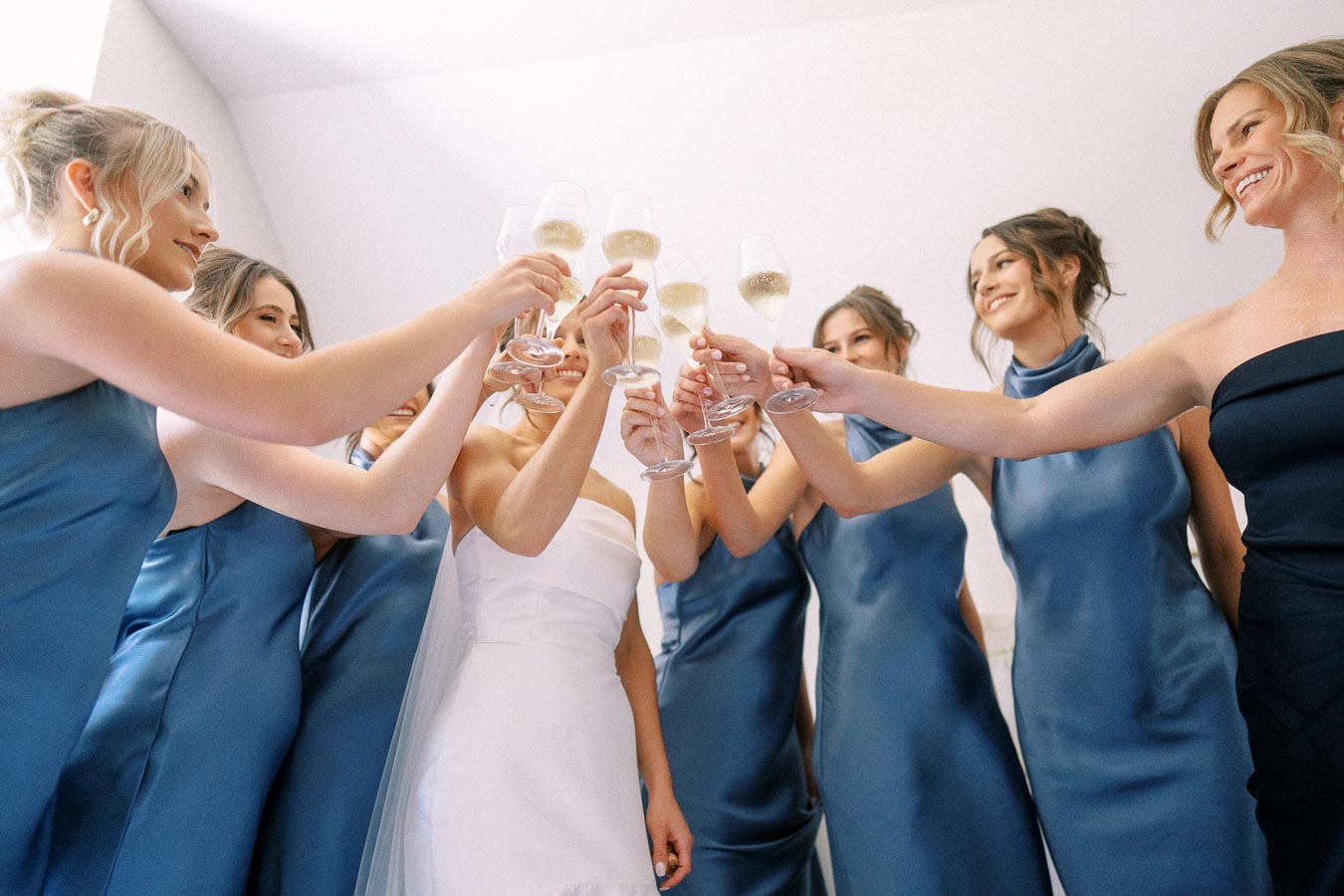 Bride and bridesmaids in blue dresses toast with champagne indoors during wedding morning preparation near Chatham-Kent, Ontario.