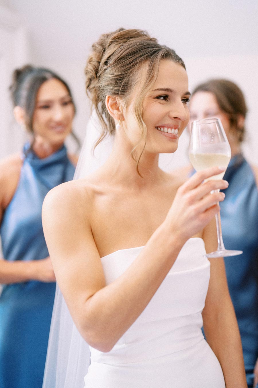 Smiling bride in a white strapless wedding dress holding a glass of white wine with bridesmaids in blue dresses behind her in Chatham-Kent, Ontario.