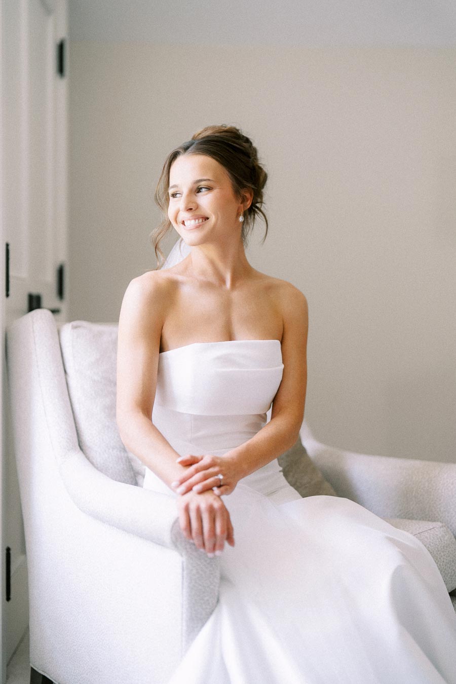 Bride in a strapless white wedding dress seated in a light armchair, smiling beside a window during bridal morning portraits.