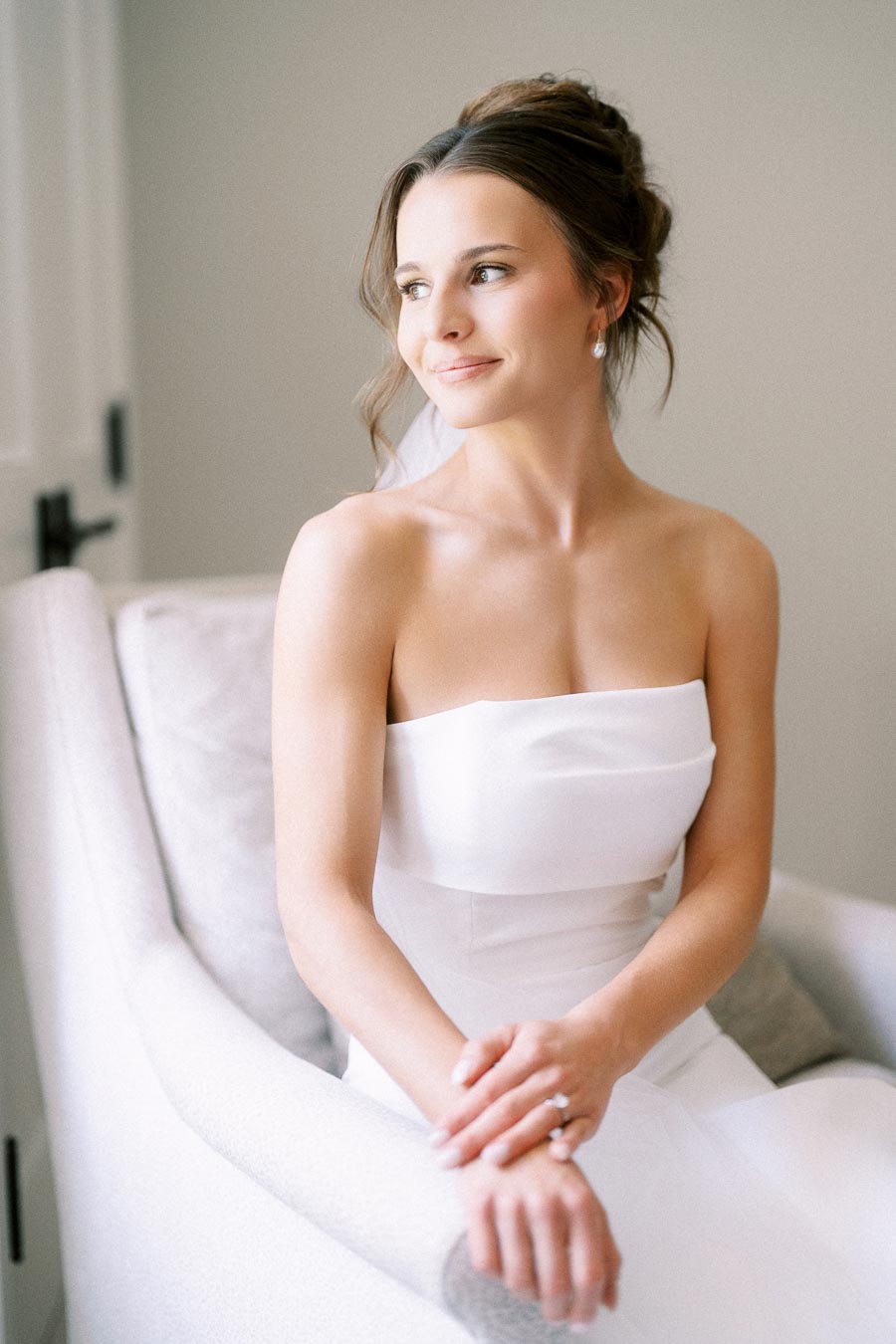 Bride in a strapless white wedding dress sitting in an armchair by soft window light during bridal morning in Ontario.