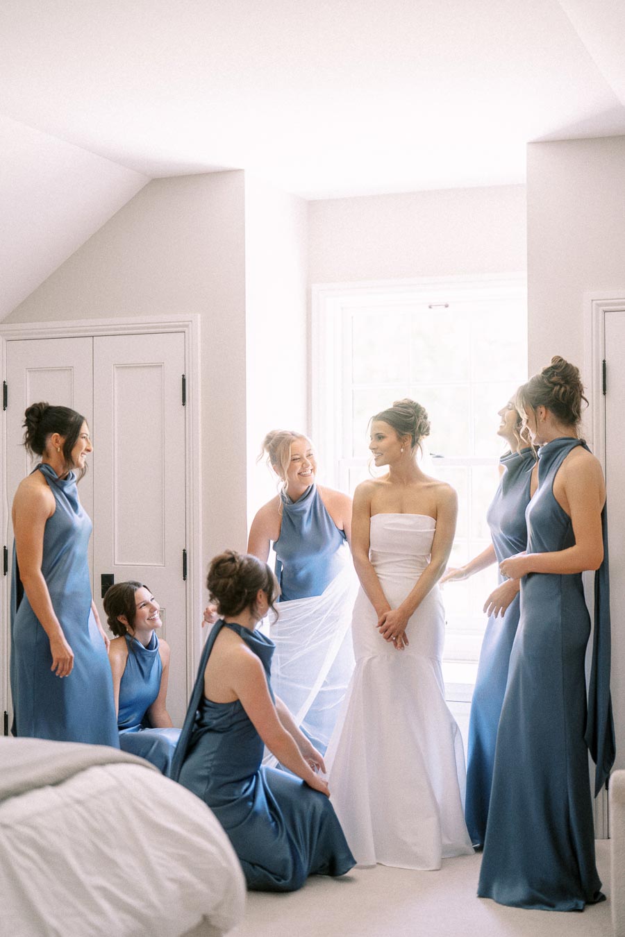 Bride in a white strapless wedding dress surrounded by bridesmaids in blue dresses inside a bright bedroom near Chatham-Kent, Ontario.
