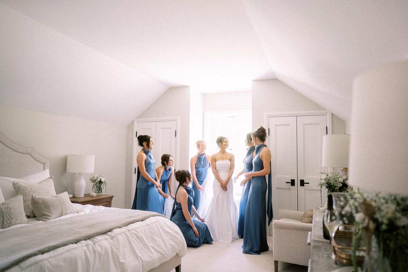 Bride in a white wedding dress stands with bridesmaids in blue dresses inside a bright attic bedroom in Chatham-Kent, Ontario.
