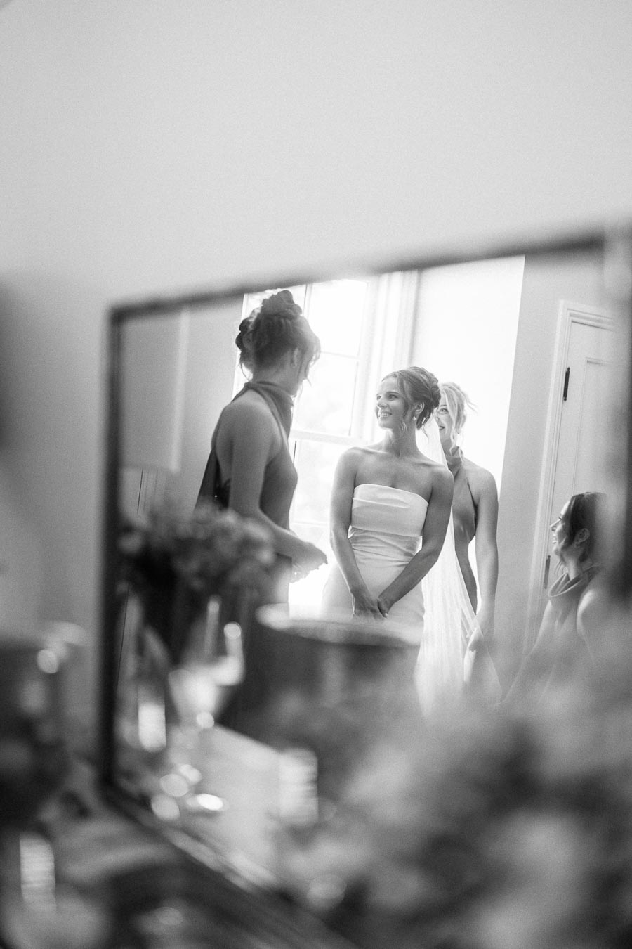 Black and white mirror reflection of a bride in a strapless wedding dress with bridesmaids during morning preparations indoors near Chatham-Kent, Ontario.