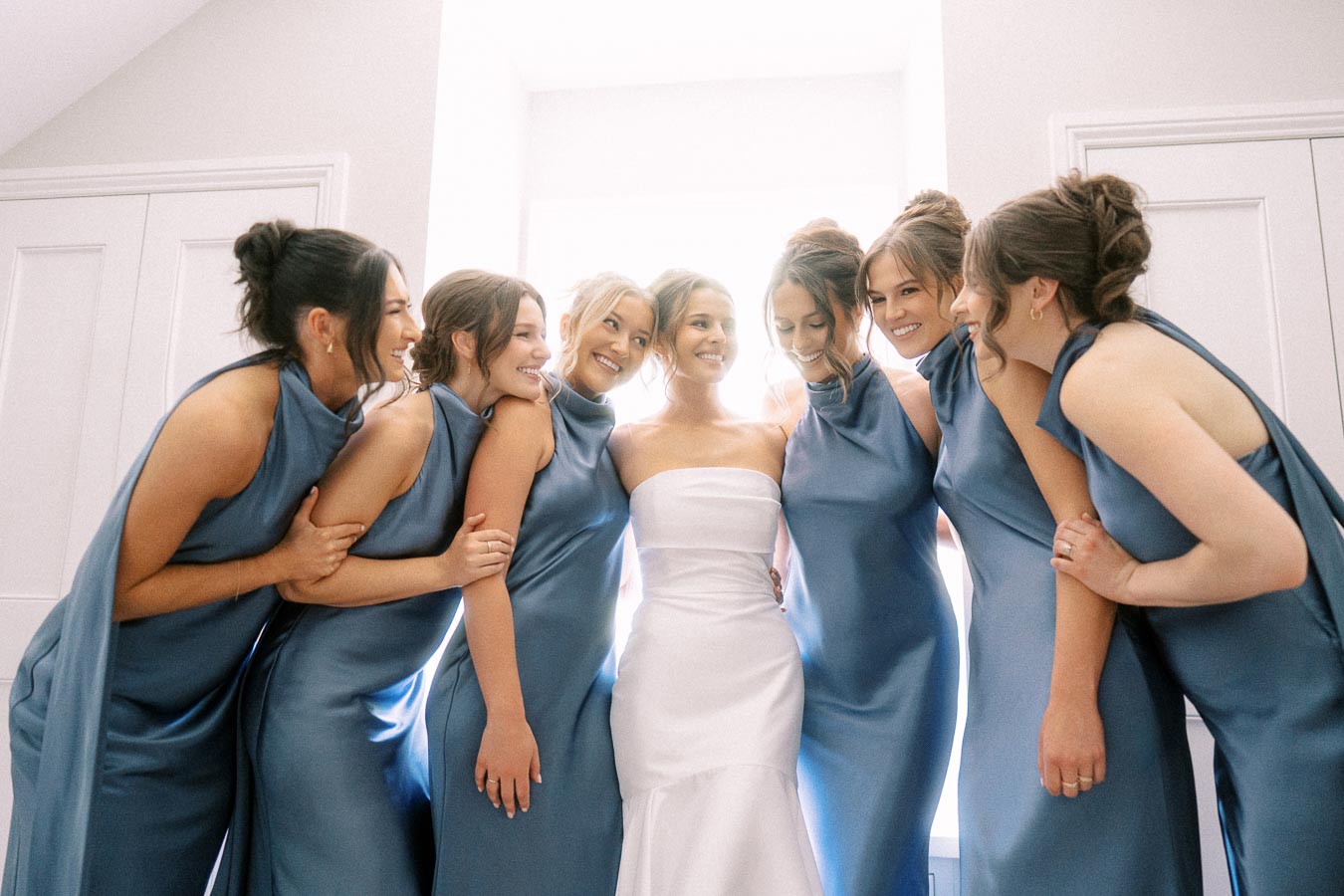 Bride in a white strapless wedding dress smiles with six bridesmaids in blue dresses during a bright indoor wedding morning in Chatham-Kent, Ontario.