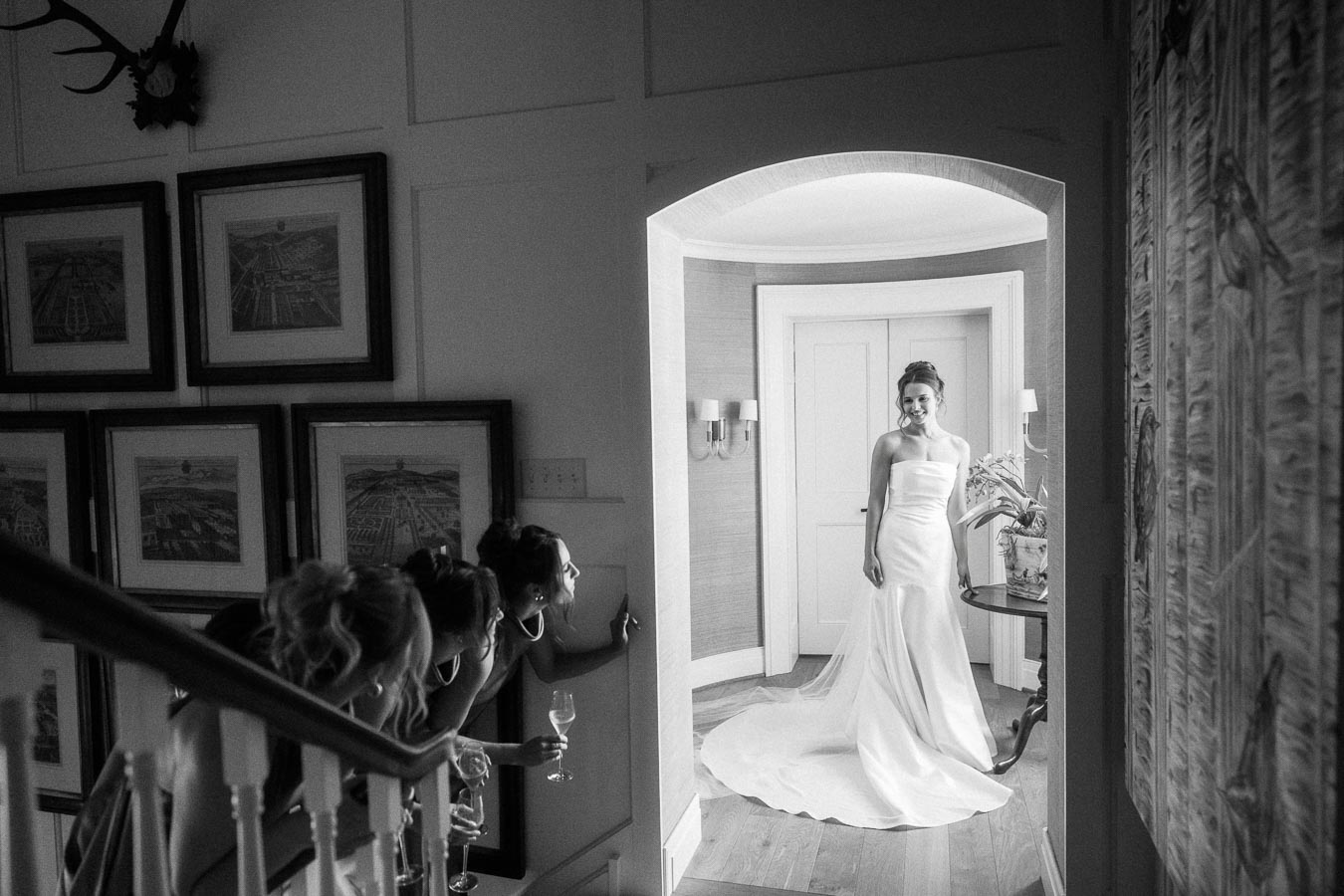 Black and white indoor wedding photo of a bride in a strapless gown standing in a doorway while bridesmaids watch from a staircase.