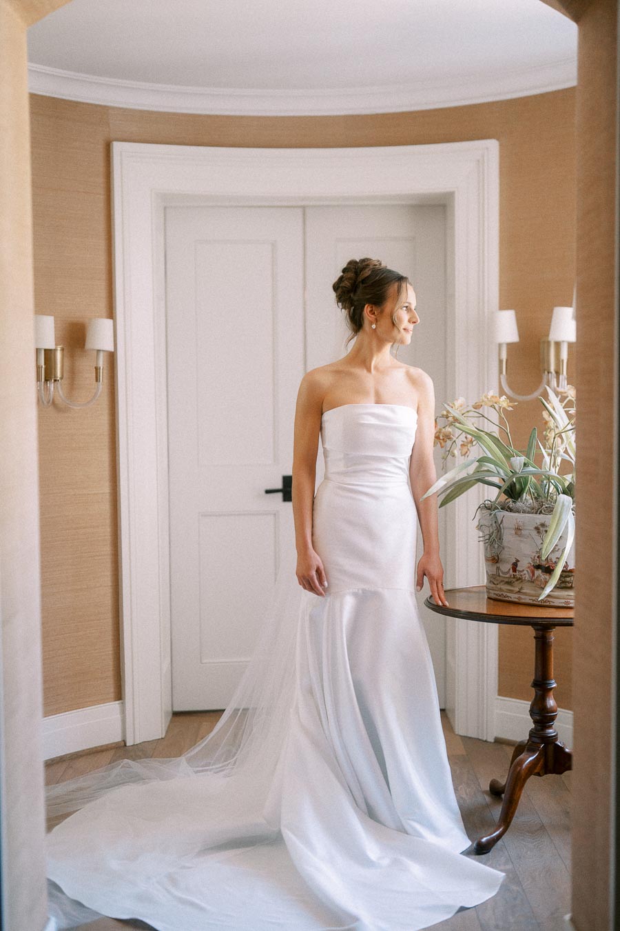 Bride in a strapless white wedding dress standing indoors beside an orchid plant on a round wooden table in Chatham-Kent, Ontario.