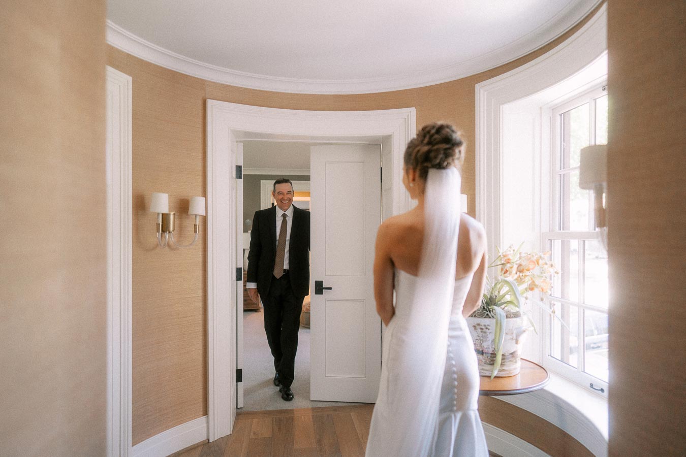 Bride in a white wedding dress and veil stands by a bright window as her smiling father enters the doorway for a first look in Ontario.