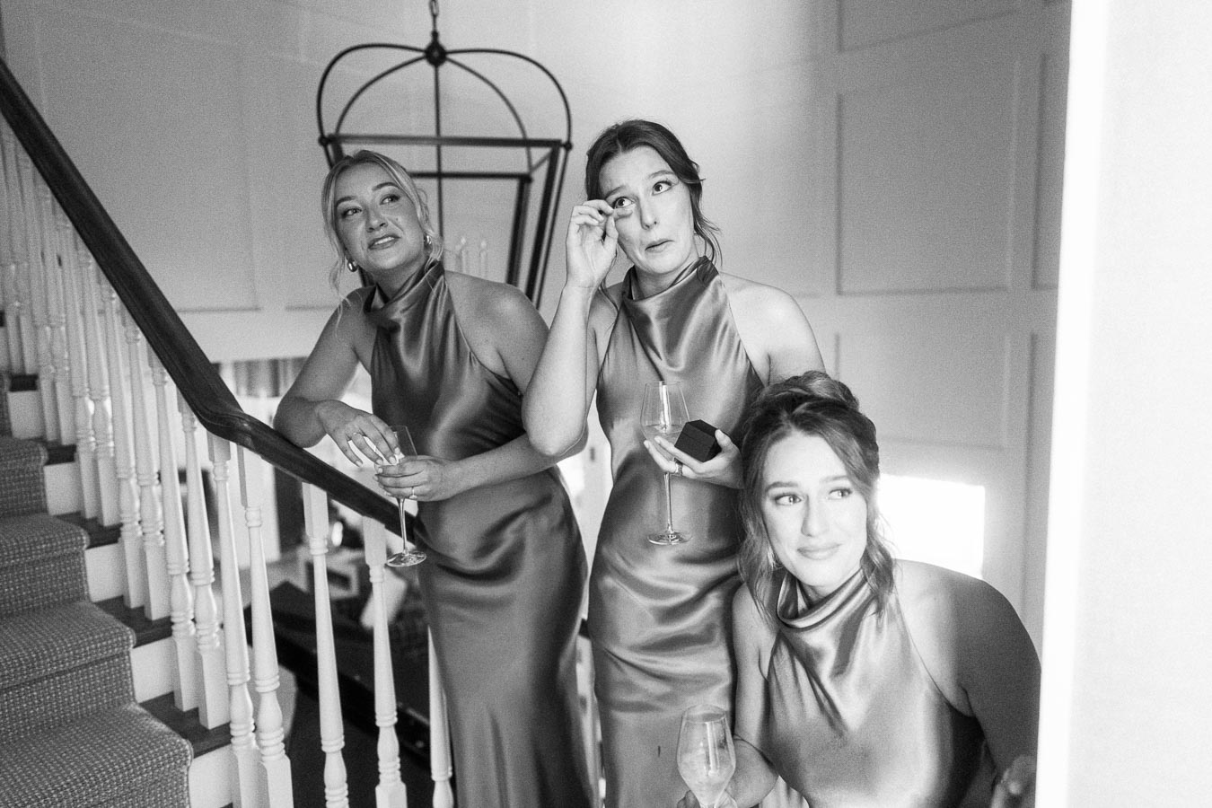 Black and white photo of three bridesmaids in satin dresses holding champagne glasses on an indoor staircase during a wedding morning in Ontario.