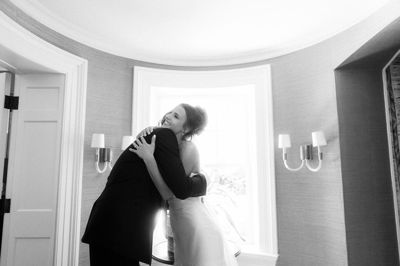Black and white photo of a bride hugging her father during a wedding morning inside a bright private estate room in Ontario.