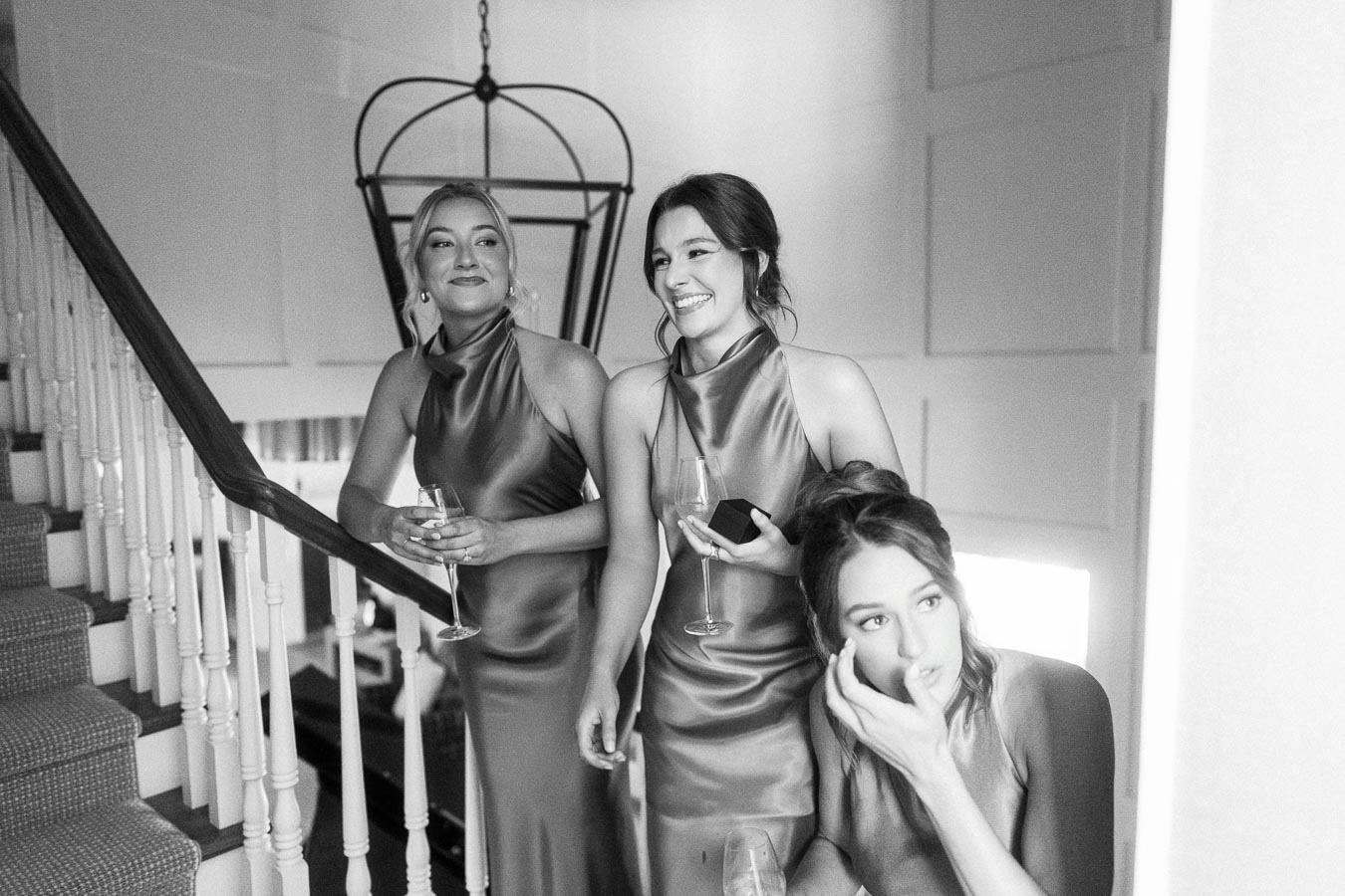 Black and white photo of three bridesmaids in satin dresses holding champagne glasses on an indoor staircase in Ontario.