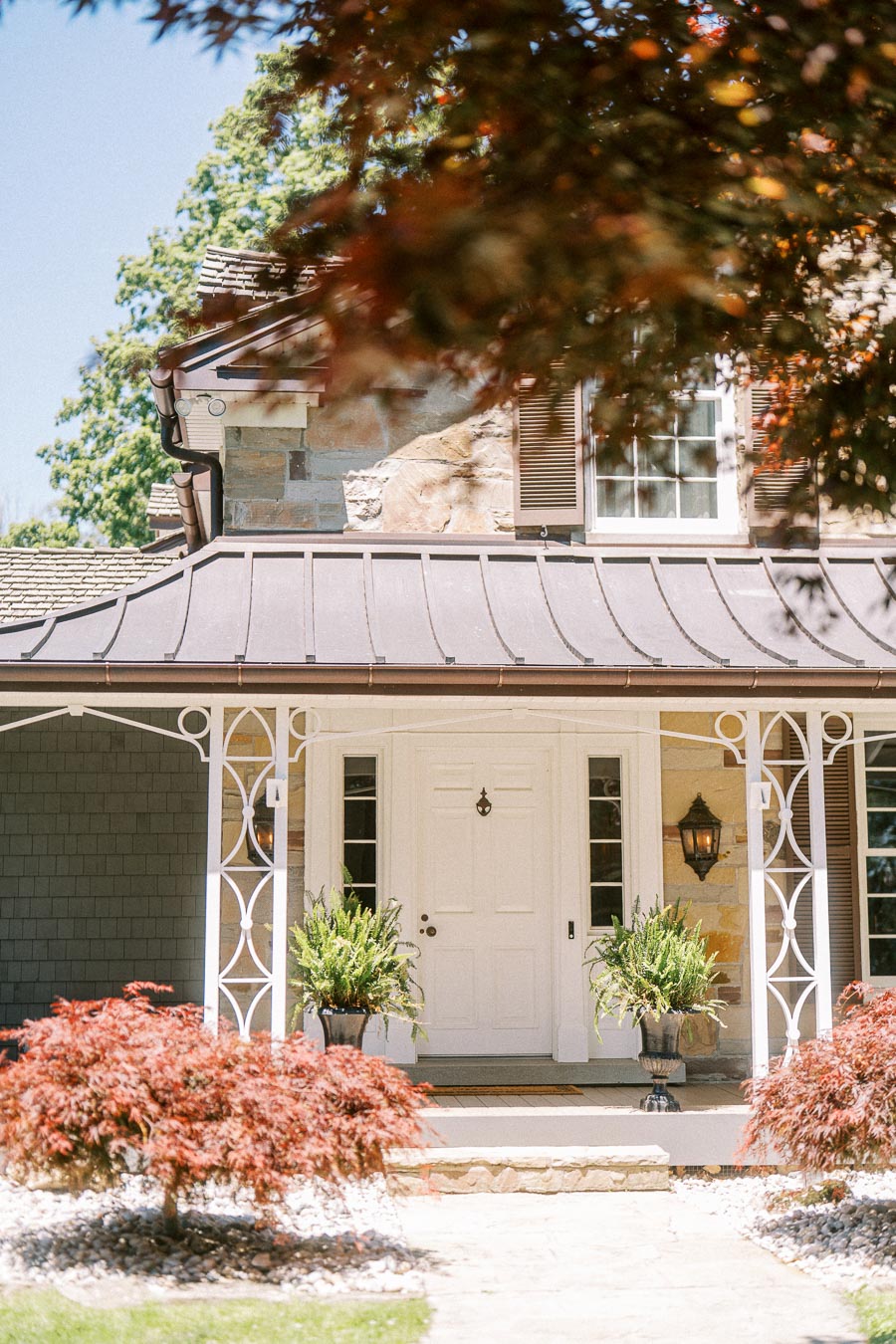 Sunlit stone house entrance with white porch columns, potted ferns, red Japanese maples, shutters, and a garden walkway in Ontario.