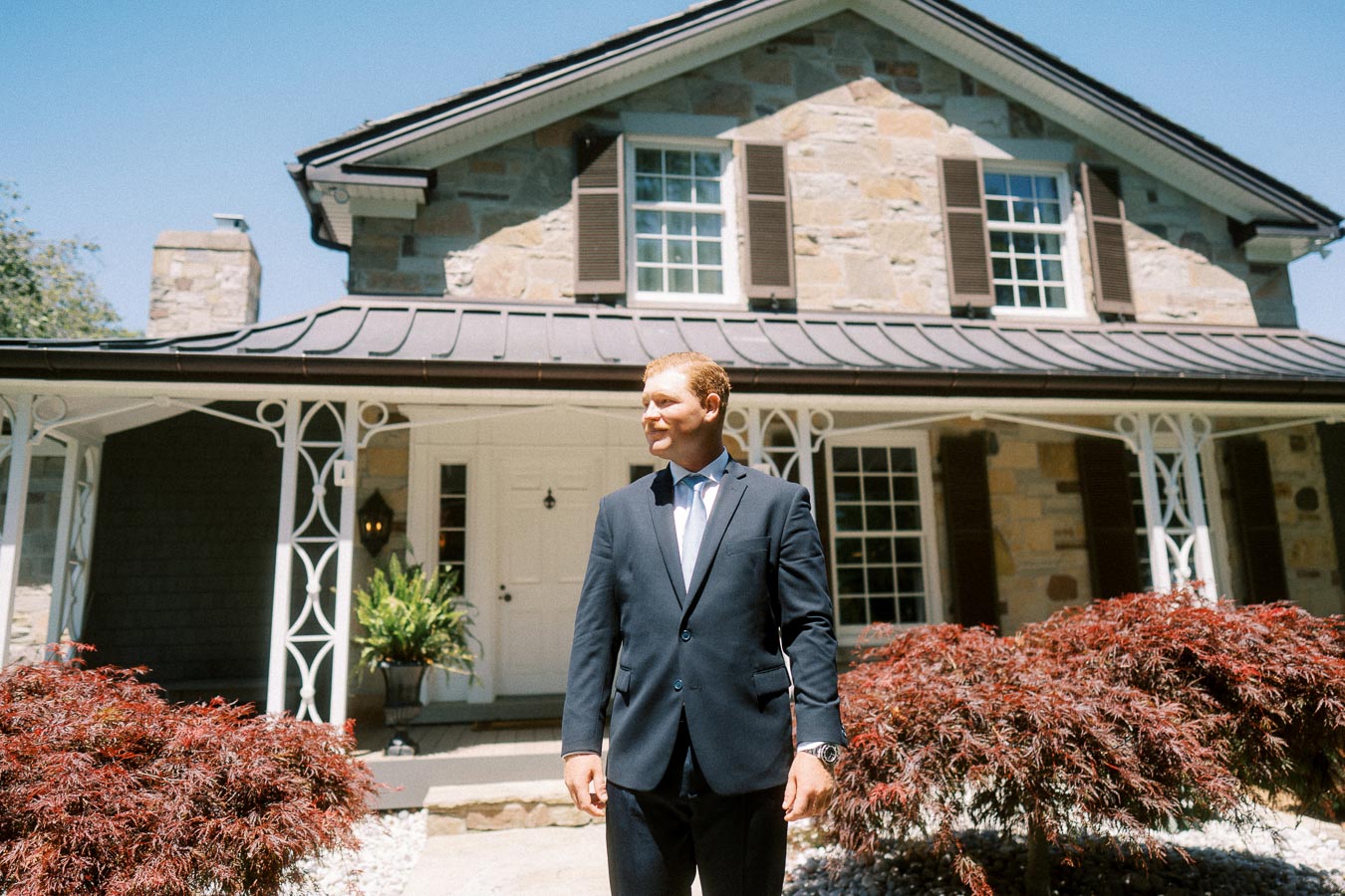 Groom in a navy suit standing outside a stone private estate house in Chatham-Kent, Ontario, on a sunny wedding day.
