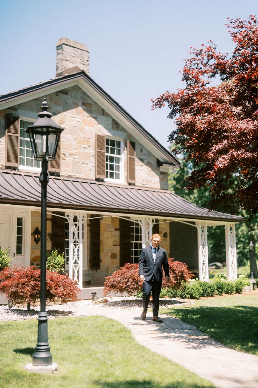 Groom in a navy suit walking along a garden path outside a stone private estate house near Chatham-Kent, Ontario.