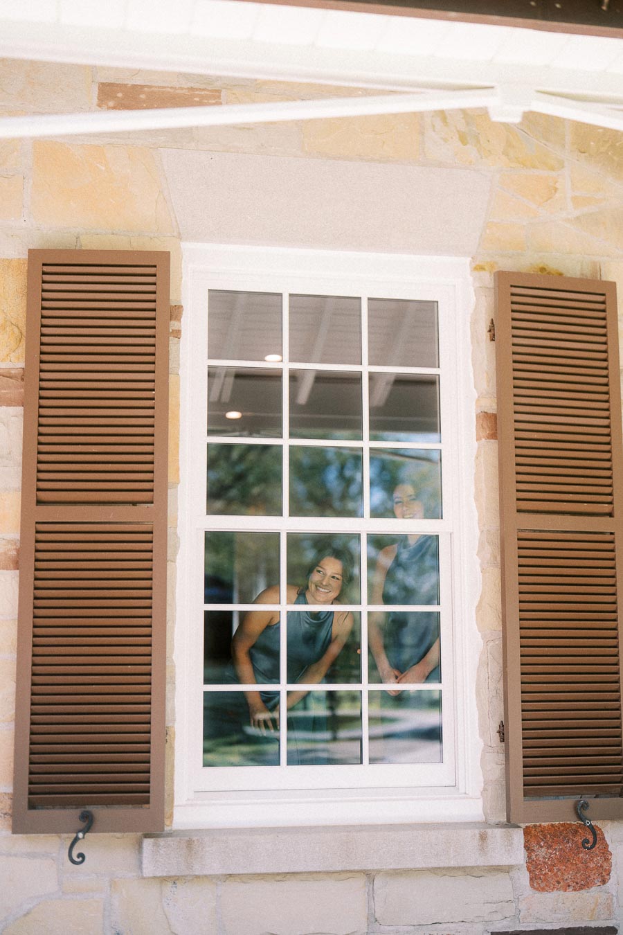 Two women look through a white framed window with brown shutters on a stone house during a private estate wedding in Ontario.