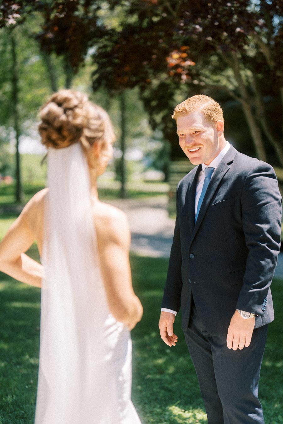 Groom smiling at bride during first look outdoors on a tree lined private estate lawn near Chatham-Kent, Ontario, Canada.